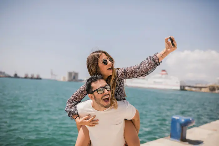 un chico cargando a su novia mientras ella toma una foto con un paisaje muy veraniego en la playa 